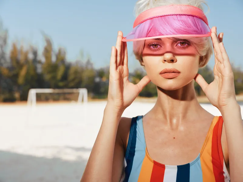 A stylish model on a sunlit beach adjusts a pink visor, wearing a bold striped swimsuit and glossy skin, creating a striking fashion moment under clear blue sky.