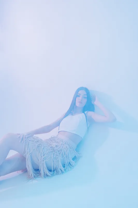 Young woman beauty fashion model lying on the floor wearing a white crop top and fringed skirt. Soft lighting creates a dreamy atmosphere. Concept of style, elegance, and modern trends.