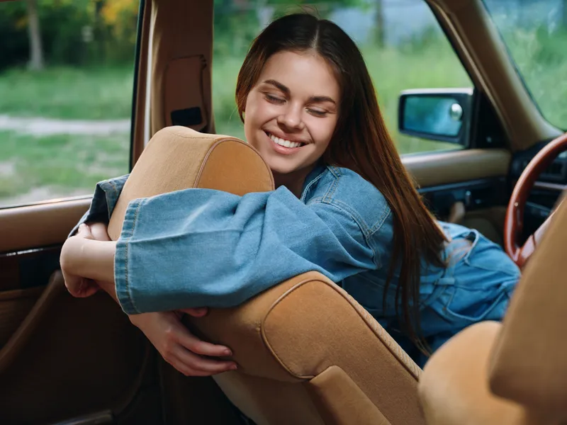 A young woman of Caucasian descent happily embraces the car seat, wearing a denim jacket, in a vibrant green outdoor setting, conveying joy and comfort.