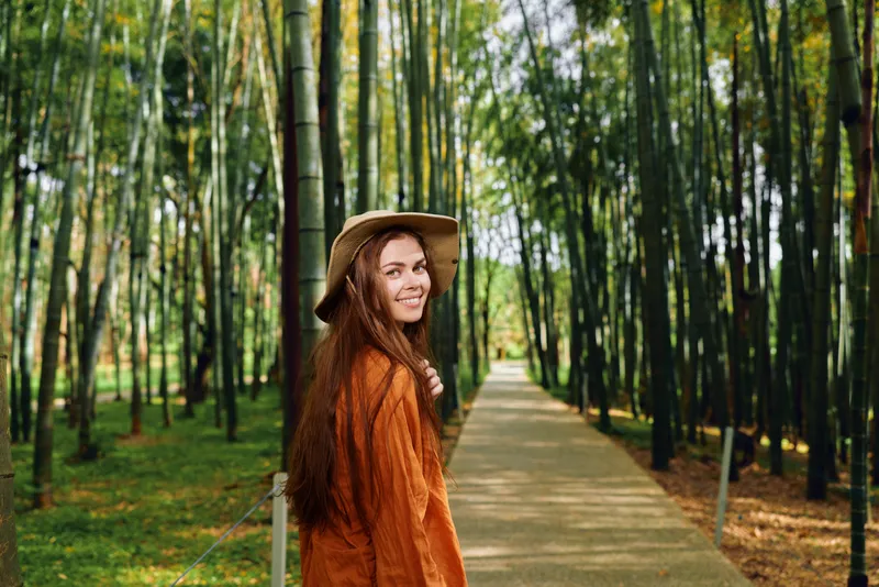 Woman with a warm smile wearing hat and coat walks along a bamboo-lined path and walkway through a forest, portrait of a traveler enjoying nature, green grove and calm outdoor mood.
