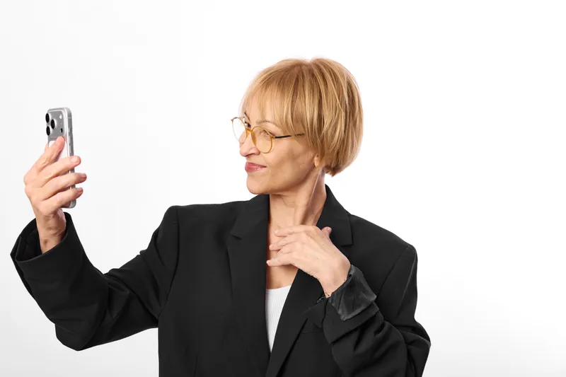 Businesswoman woman takes selfie with phone smartphone, glasses, studio portrait, hand on neck in thinking pose, professional calm expression on white background