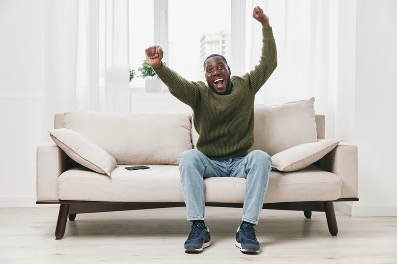 Happy African American man celebrating success while sitting on a cozy sofa, wearing a casual green sweater and jeans in a bright, modern living room