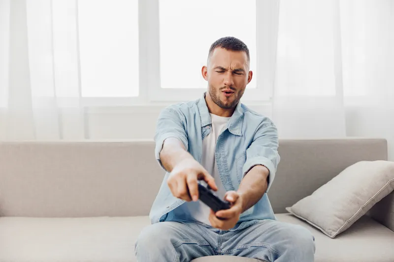 A focused man enjoying video gaming at home, wearing a casual blue shirt. The bright room enhances the excitement of his gaming experience, showcasing modern leisure.