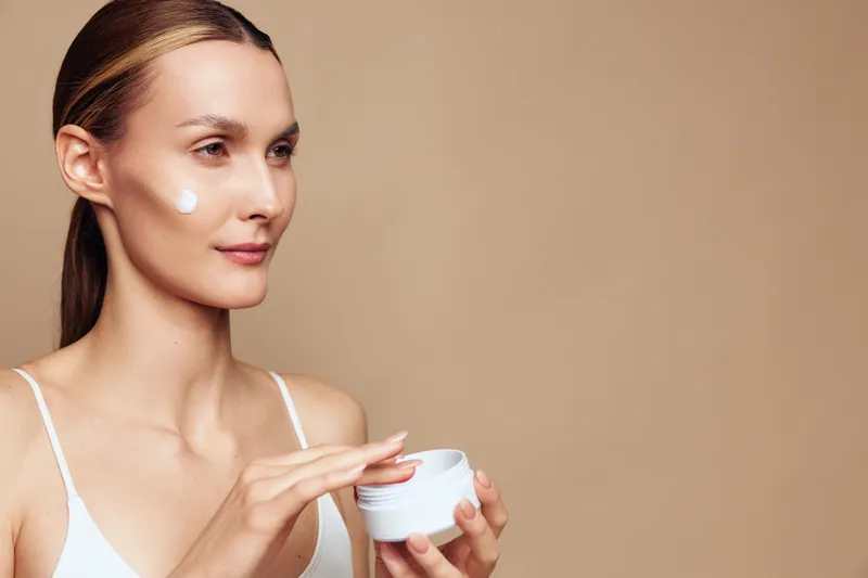 A woman applies moisturizer to her cheek in a neutral studio, smiling softly. A skincare routine theme focusing on self-care, beauty, and calm daily rituals.