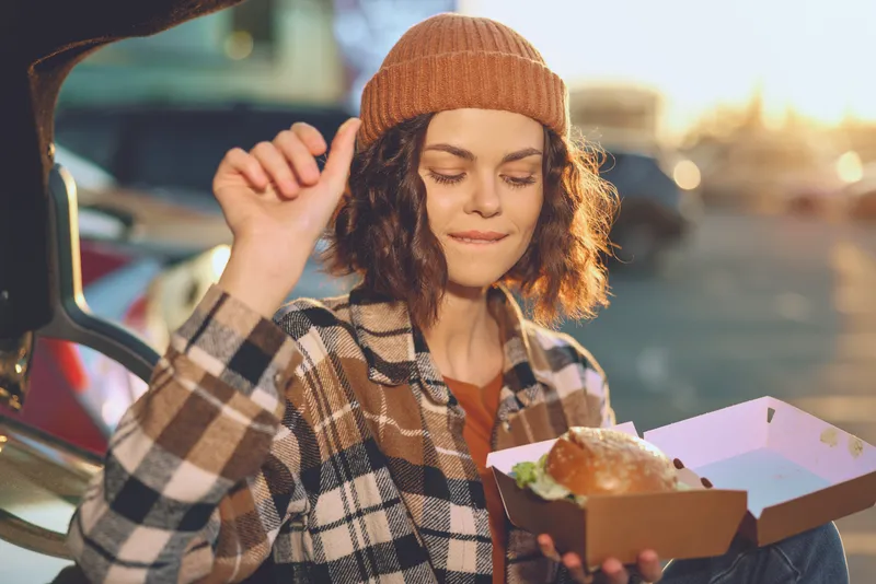 woman enjoying a burger from a takeaway box wearing a beanie and plaid jacket during golden hour glow, candid urban portrait conveying authenticity, mindful living and emotional storytelling.