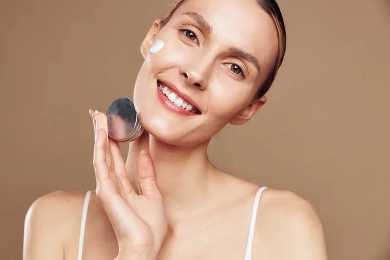 Happy woman applies cream on cheek with a spatula, showcasing skincare routine, smiling at camera, shoulders exposed, beige background, skin care concept, beauty ritual