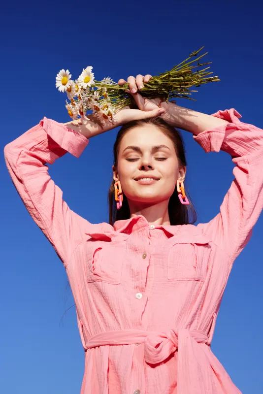 Young woman in a pink dress holding flowers against a blue sky, radiating joy and positivity in a vibrant outdoor setting