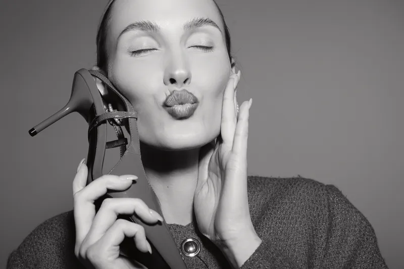 Caucasian woman with light skin holds high heel shoe near face and makes kiss expression in studio. Concept of fashion and beauty.