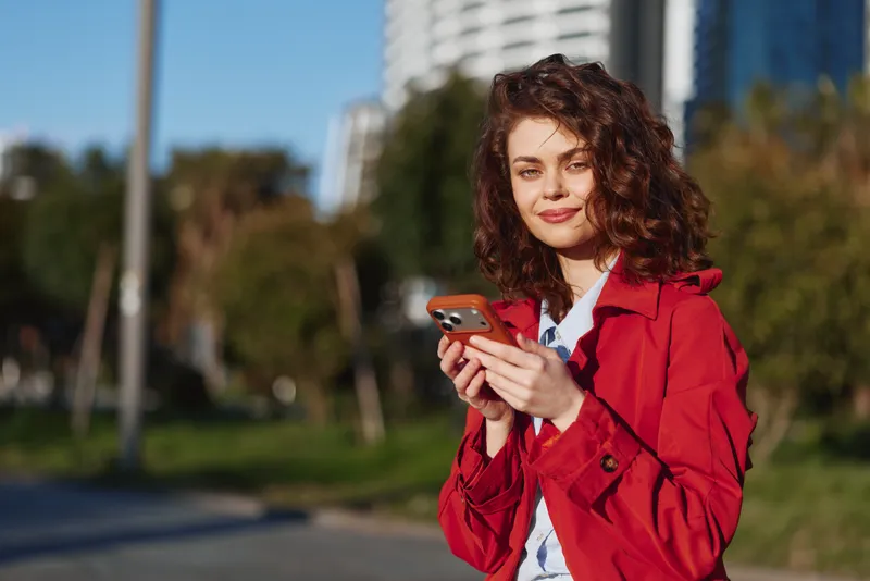Cheerful woman with curly hair wears a red jacket, smiles, and looks at a smartphone outdoors in an urban park setting, capturing a candid everyday moment