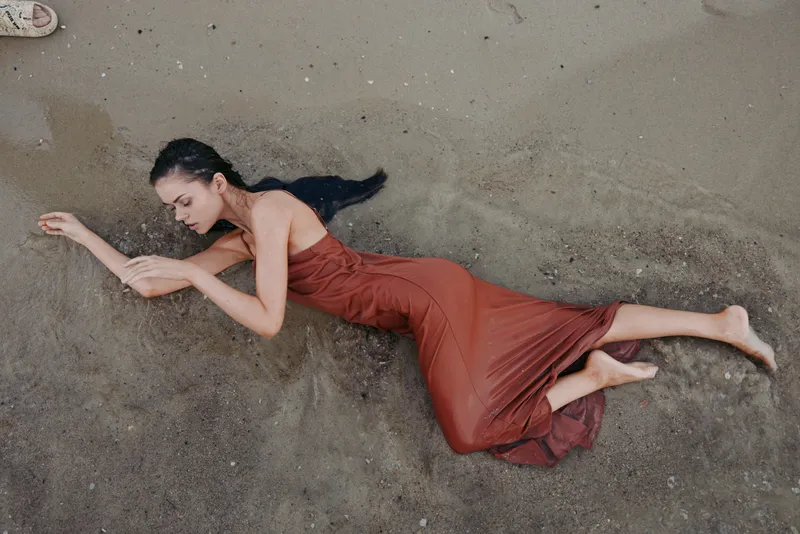 Young woman lying on wet sand wearing a brown dress with a peaceful expression and long dark hair, natural beach setting, calm and contemplative mood.