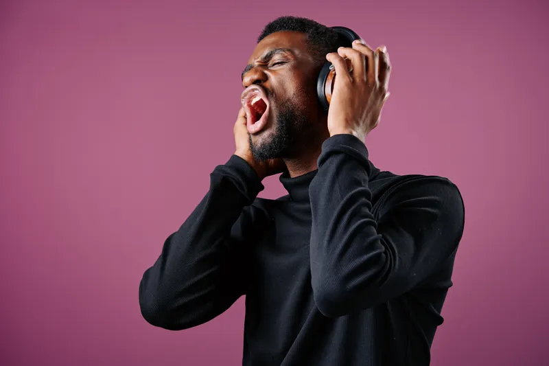 A young Black man expressing strong emotions while wearing headphones against a pink background, capturing the passion of music and self expression