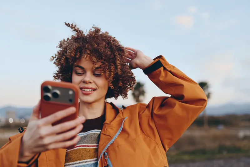 Woman lifestyle outdoors, selfie with phone, curly hair woman smiling, orange jacket, holding smartphone near face, golden daylight with soft clouds, distant hills and sky background, relaxed casual