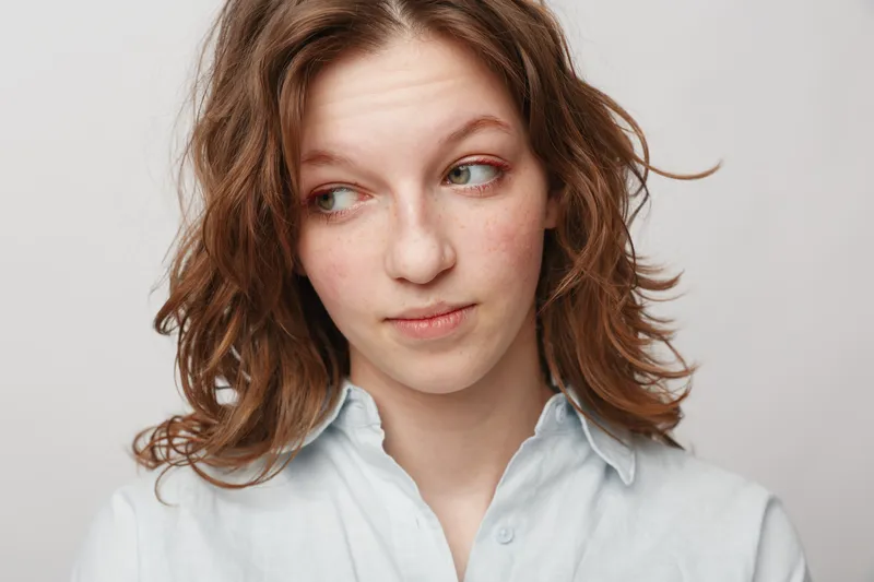 Young woman with curly hair looks sideways with thoughtful expression wearing light blue shirt on plain background. Concept for contemplation, emotion, and casual portrait.