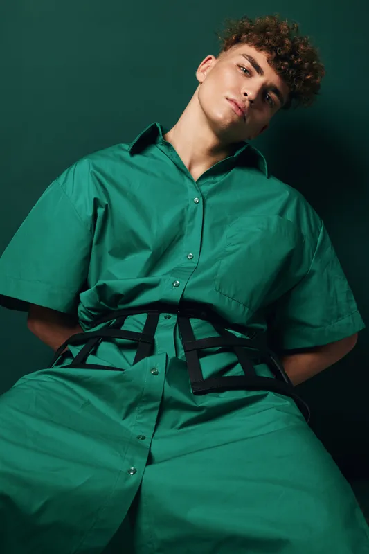 Young man with curly hair wearing oversized green shirt and pants posing against dark green background. Fashion model with casual style and confident expression in studio portrait.