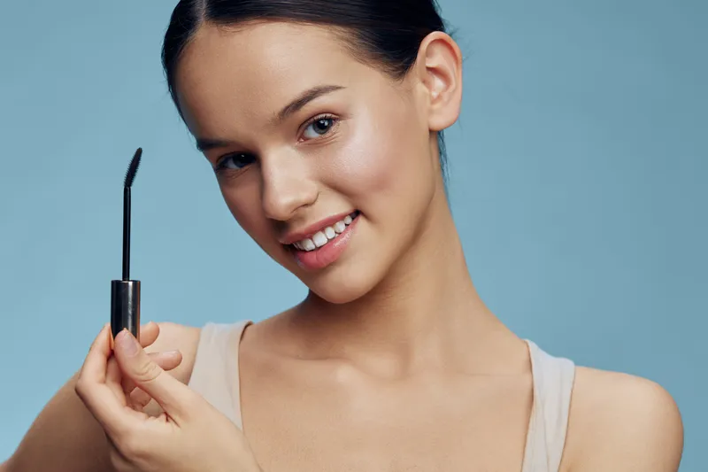 Young woman with flawless skin holding mascara, showcasing beauty and makeup trends against a soft blue background, exuding confidence and joy