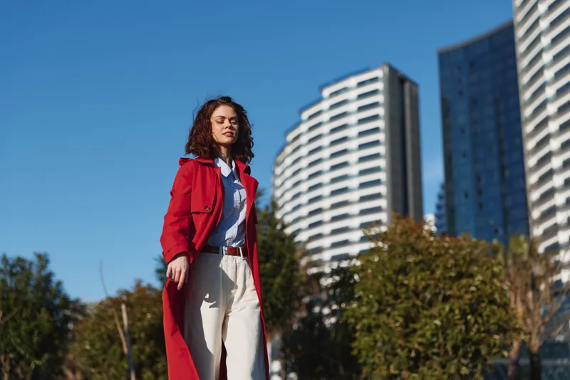 Young woman in red coat walking outdoors in city, sunny day, modern buildings background, confident expression, casual style, natural light.