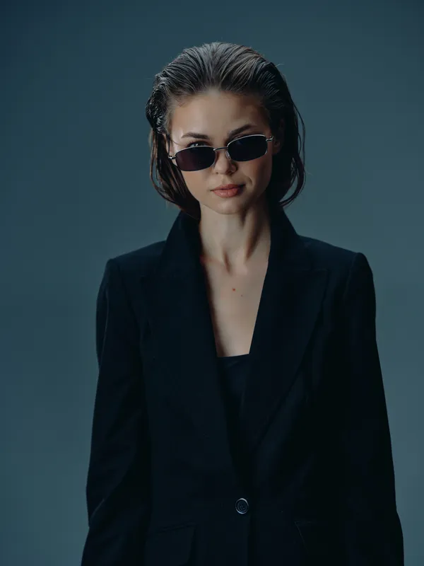 Stylish woman wearing sunglasses and a black blazer stands confidently against a dark background with a moody atmosphere.