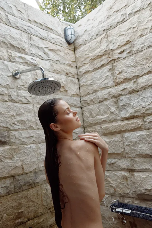 naked woman showering outdoors under rain showerhead with wet long hair against textured stone wall in natural light setting