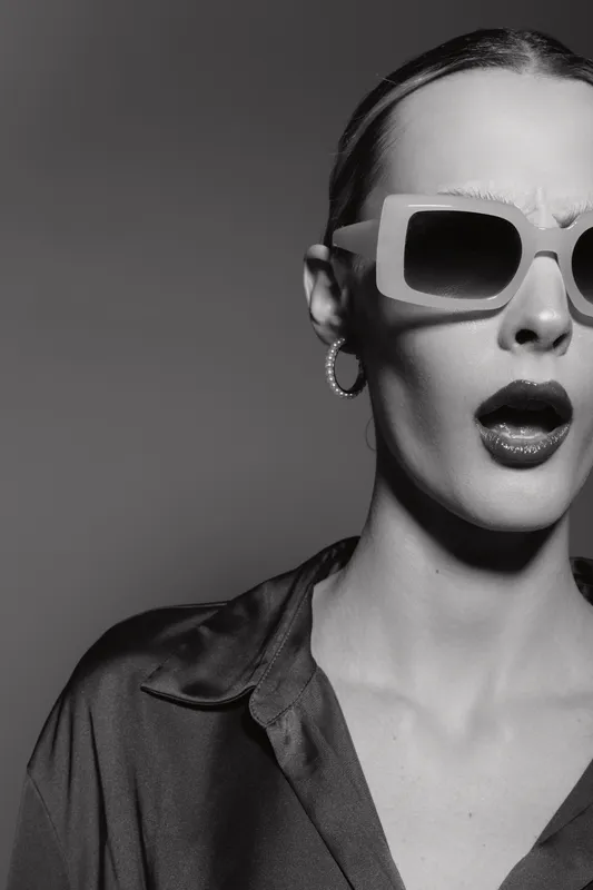 Fashionable white woman with pale skin wearing large sunglasses and silk shirt looks surprised in black and white studio portrait.