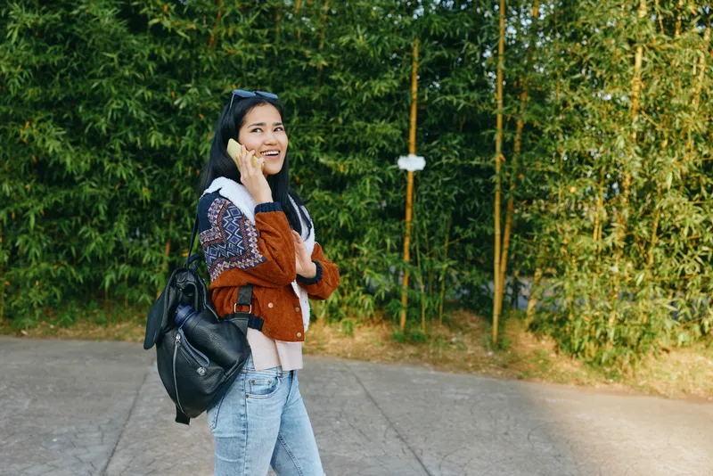 Woman on a phone call outdoors with a backpack, casual sweater and jeans, smiling as she chats on a sunny path lined with trees during daytime.