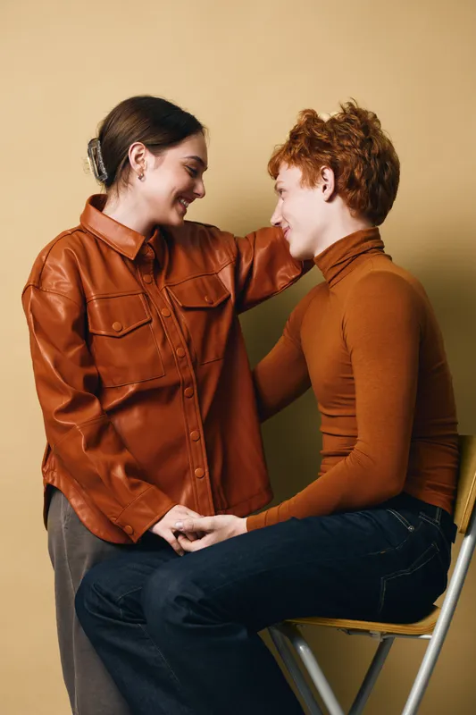 Couple sitting and smiling with gentle touch showing affection and connection, two people in casual warm clothing in intimate moment on beige background.