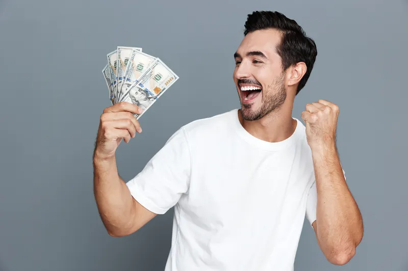 Excited smiling man in casual white t-shirt holding cash display expressing joy and triumph, isolated on solid background. Success and happiness concept