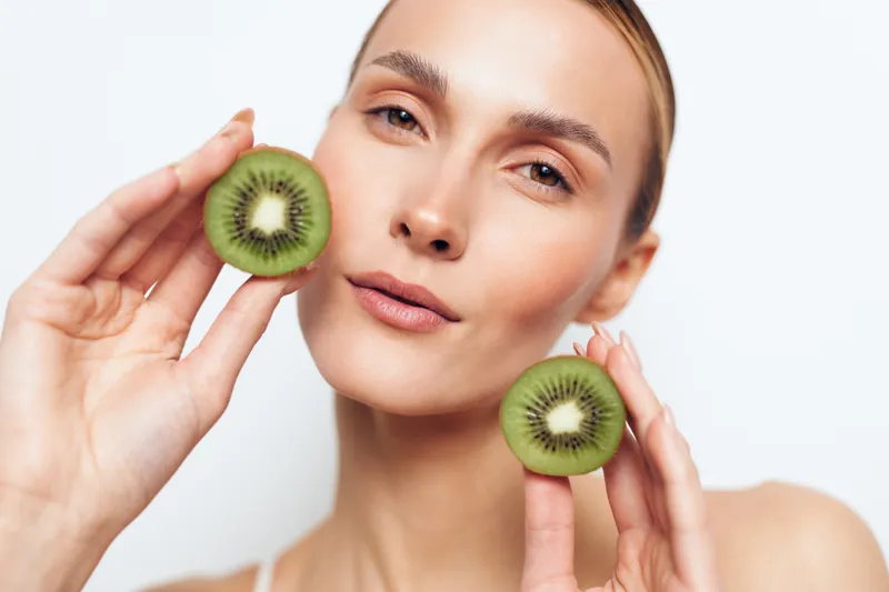 Woman with kiwi slices at her face in close up on a clean studio white background, fresh fruit beauty and skincare for natural wellness self care, soft lighting, calm expression