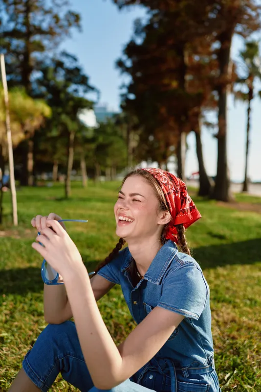 smiling woman in denim outfit laughing outdoors with red headscarf in sunny park surrounded by green grass and tall trees on a clear day