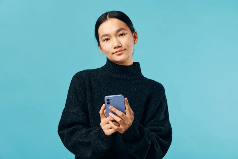 Young woman with smartphone in black sweater standing and looking at camera on blue background. Casual female holding mobile device for communication and social media.