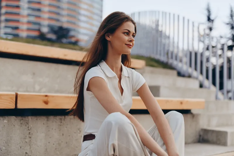 Young woman in white casual outfit sitting outdoors with confident smile in urban park setting, natural light, summer mood, casual fashion, lifestyle concept