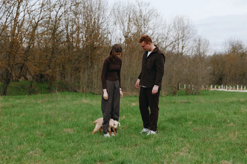 Casual couple outdoors with dog in a green field, relaxed interaction, natural colors, soft breeze, candid moment, open space, calm atmosphere, simple outfits, wholesome lifestyle