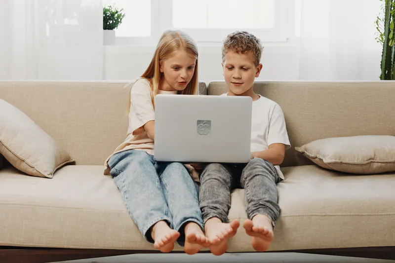 Two children sitting barefoot on couch using laptop together in bright living room. Concept of technology, learning, and childhood collaboration.