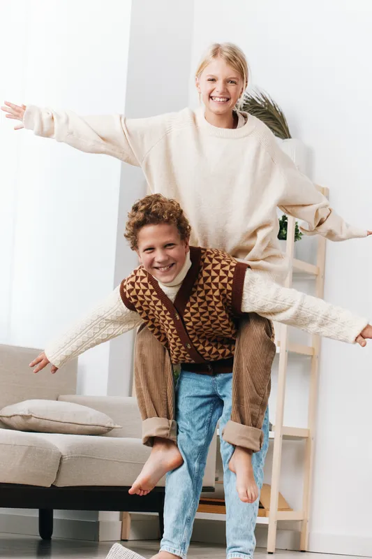 Two children share a joyful piggyback moment indoors, arms outstretched in a bright, cozy living space, capturing carefree energy and playful trust between siblings.