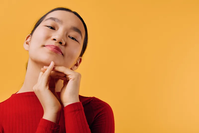 Young woman with smooth skin and dark hair wearing red long sleeve top posing with hands under chin against orange background. Calm face and confident expression in studio lighting.