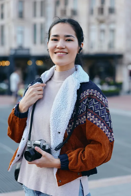 Camera, photography, woman, outdoor, street, jacket, smiling in casual urban style while holding a camera in a busy city square during daytime