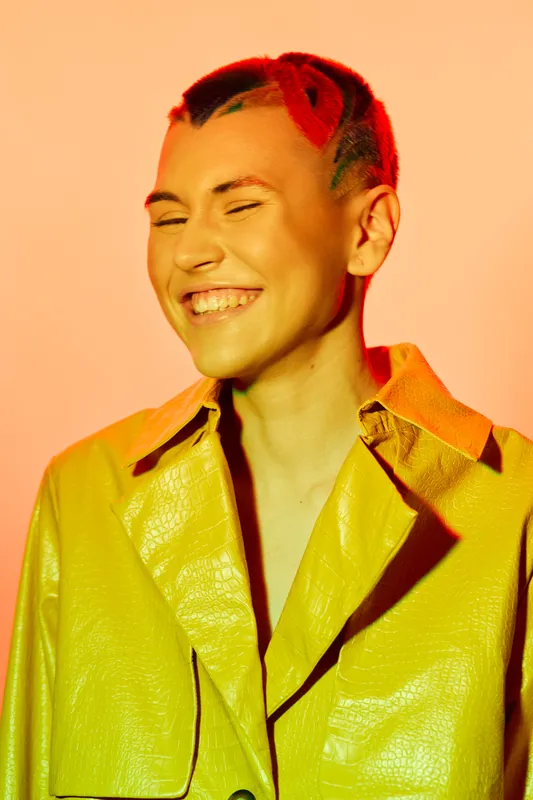 smiling woman with colorful hair wearing a bright yellow jacket, expressive joyful face, warm orange background, modern fashion style, vibrant lighting