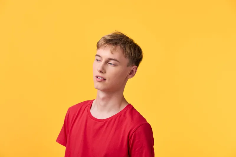 Young man in a red shirt stands against a vivid yellow background, presenting casual style and a thoughtful expression in a bright studio, with clean composition and confident vibe for lifestyle and