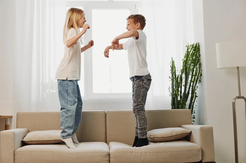 Two happy children jumping on sofa in bright living room with natural light, enjoying playful moment and sibling fun at home during daytime.
