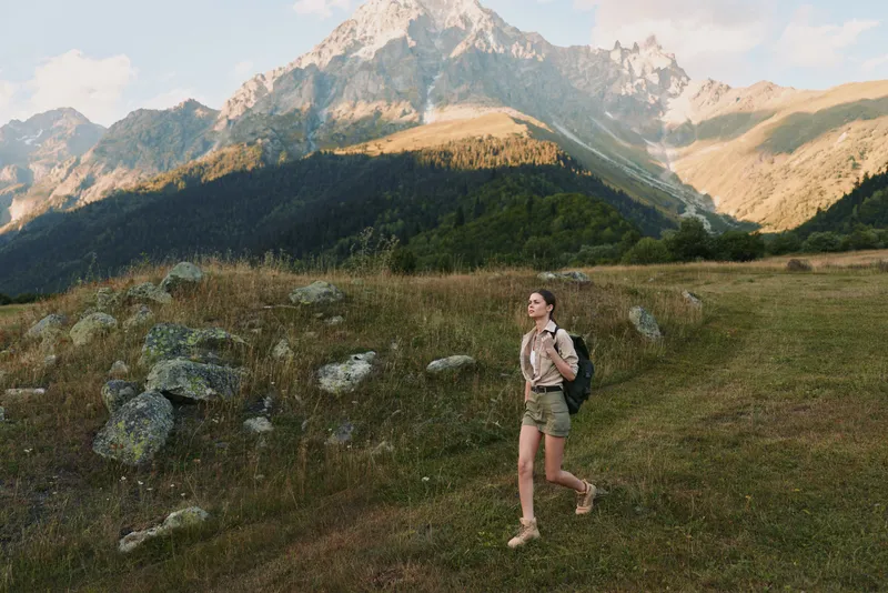 Young woman with backpack hikes through a grassy mountain valley on a sunny day, enjoying nature and outdoor adventure in casual summer clothes.