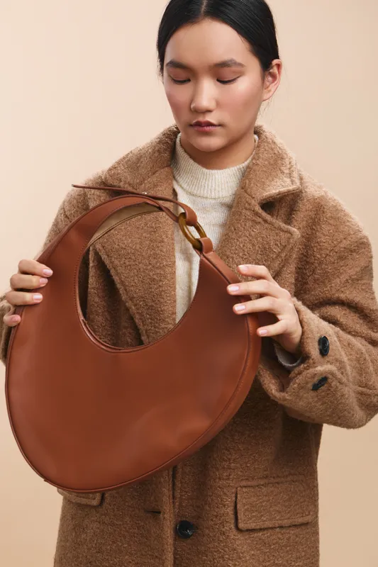 Asian woman wearing a wool coat holds a large brown circular handbag, posing in a neutral studio. Her calm expression and soft lighting create a chic, minimalist fashion moment.