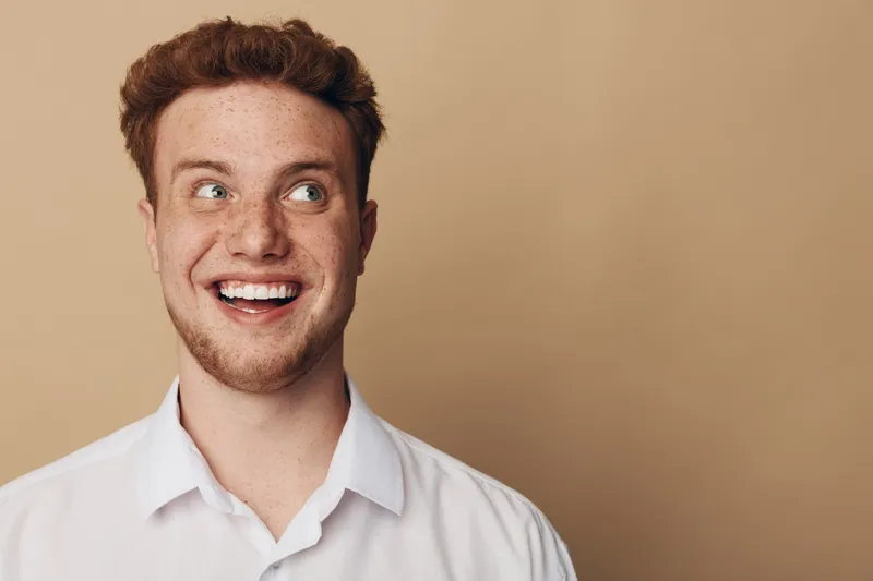 Smiling young man with red hair and freckles looks to the side wearing a white shirt. Portrait on beige background with natural expression and casual style. Concept of happiness and youth.