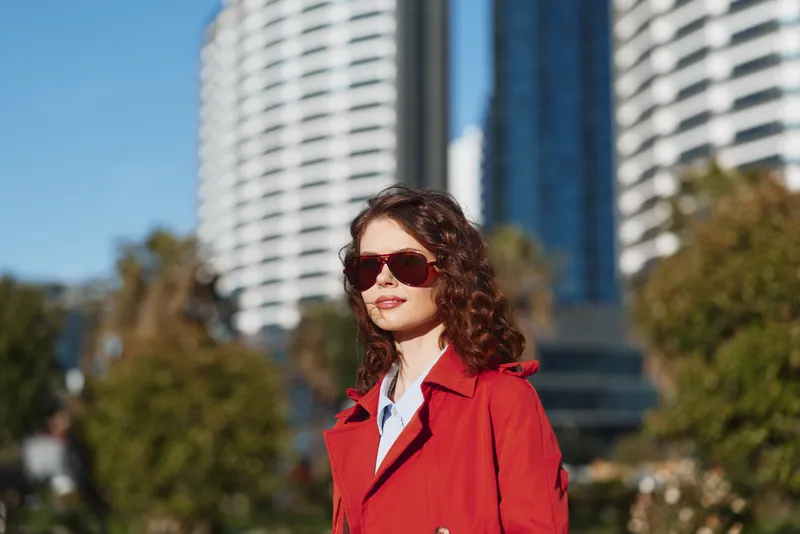 Young woman with curly hair wearing a red coat and sunglasses walks confidently in an urban park with modern buildings in the background on a sunny day.