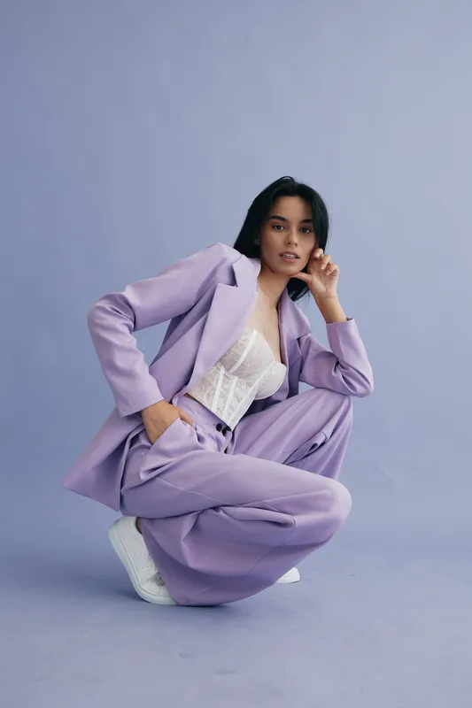 Young woman in lilac suit posing confidently against purple background, wearing white lace top and sneakers for a modern stylish look in fashion photography.