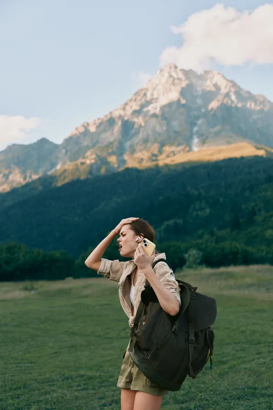 A young woman in casual clothes stands in a green field with a large backpack, touching her hair while looking away. Mountains rise in the background under a partly cloudy sky.