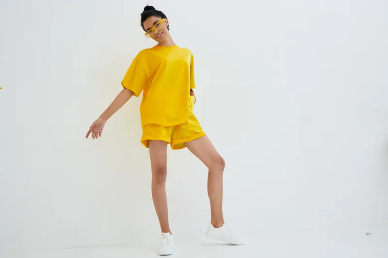 Young woman in yellow casual outfit posing confidently against white background, wearing bright sunglasses and white sneakers, showcasing vibrant summer style.
