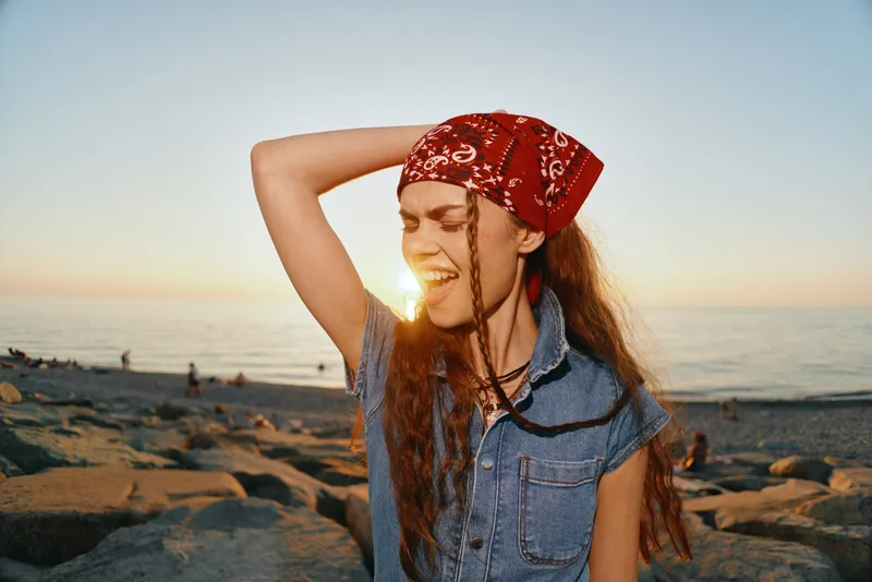 lifestyle woman in modern boho-western denim outfit with red bandana enjoys golden hour on rocky beach, film color tone highlighting relaxed street style vibe
