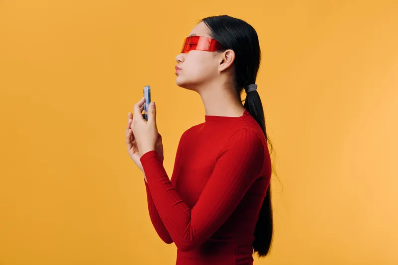 Young woman in red dress and red sunglasses with sleek ponytail holding perfume bottle, posing on yellow background for fashion beauty and fragrance concept.