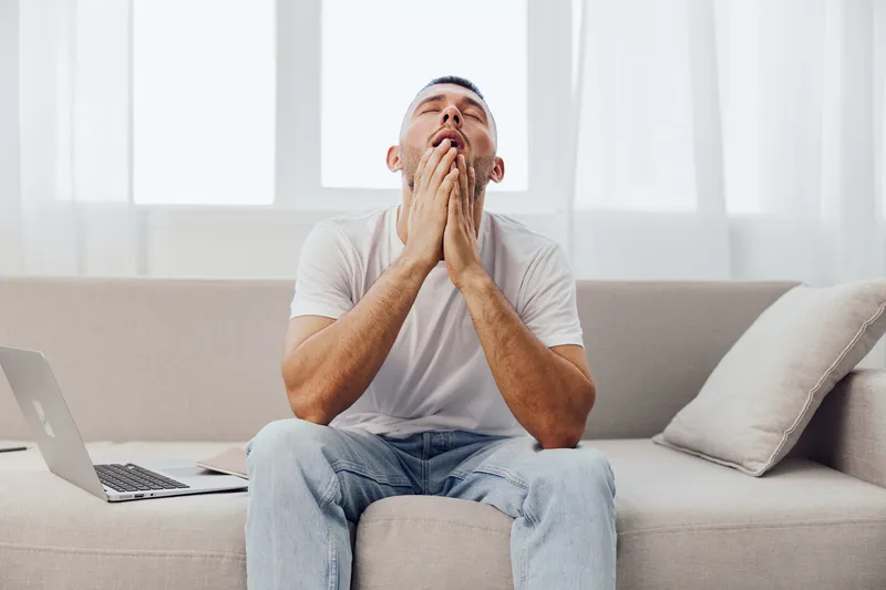 Exhausted man sitting on the sofa, showing frustration, working from home. Modern workspace with laptop, bright sunlight creates an uplifting atmosphere.