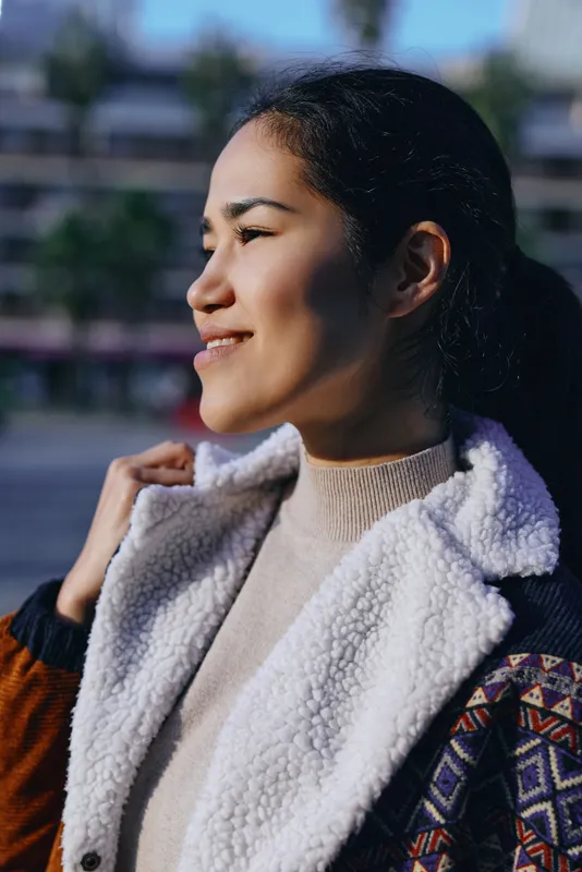 Woman with a warm smile in outdoor light, wearing a fleece-lined jacket and cozy sweater, casual street style shot capturing relaxed confidence and natural texture and color.