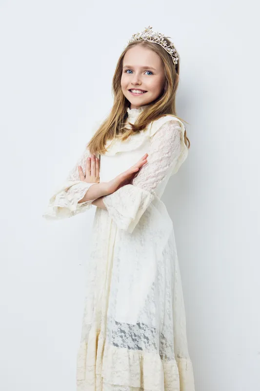 Happy young girl in a white lace dress and crown, posing confidently against a light background, ideal for themes of childhood and innocence